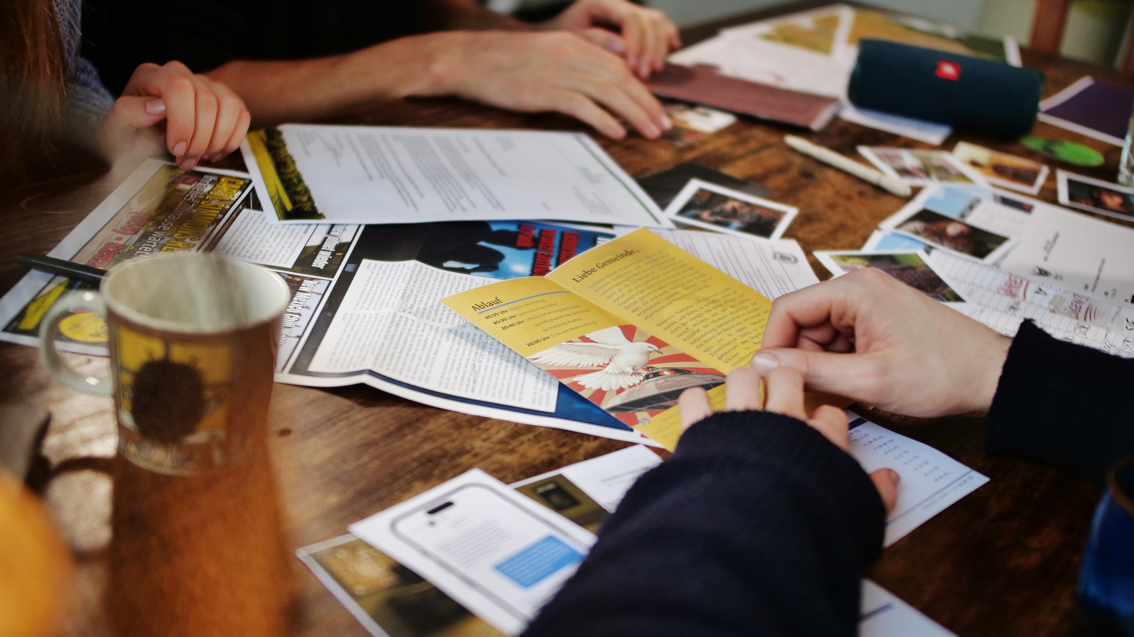 Blick auf einen Tisch, auf dem das Spielmaterial verstreut liegt: Flyer, Zeitungen, Fotos, Kalender, Berichte, Screenshots. Drei Personen sitzen darum herum und durchsuchen die Materialien, man sieht nur ihre Hände. Im Hintergrund ist eine Musikbox zu sehen, mit der die Audio-Inhalte abgespielt werden.